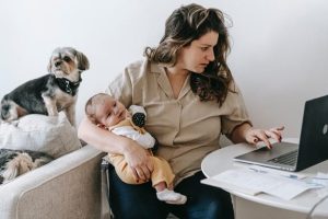 parent holding a baby while typing on an open laptop with a smal dog behind her on a couch 
